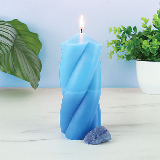 Blue twisted candle with a flame, surrounded by green plants on a light surface.