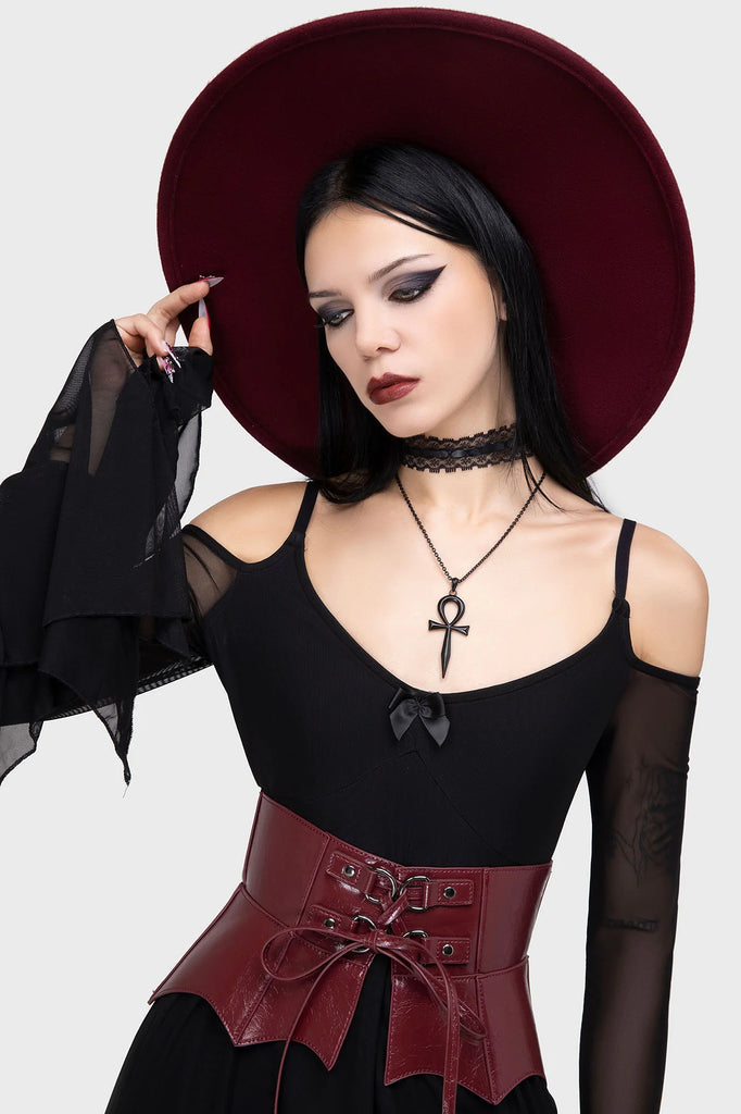 Woman in gothic attire with a maroon hat and corset, wearing a necklace with an ankh pendant.