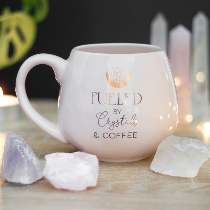 White mug with 'Fueled by Crystals & Coffee' text on a wooden surface with crystals and candles.