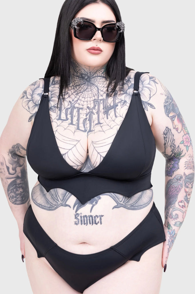 Person wearing a black swimsuit with tattoos visible on arms and chest against a plain background
