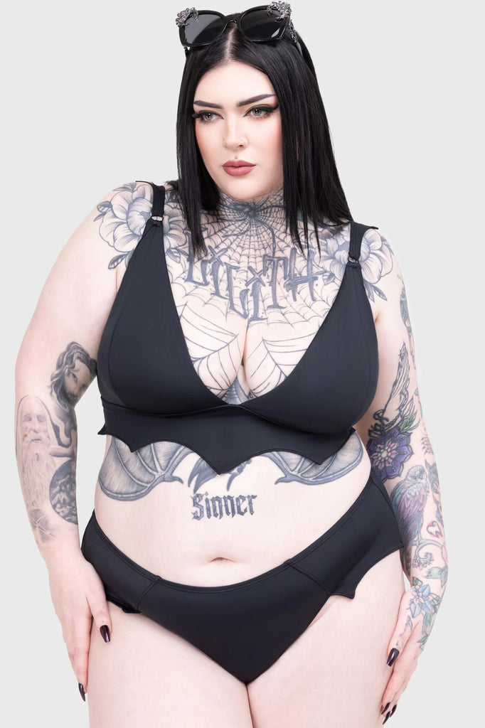 Person wearing a black bikini with tattoos on arms and chest against a light gray background