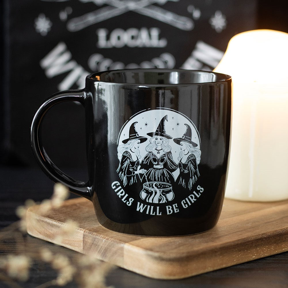 Black mug with witch design and 'Girls Will Be Girls' text on a wooden surface.