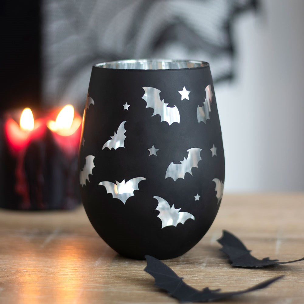 Black wine glass with bat and star designs on a wooden table with blurred candles in the background.