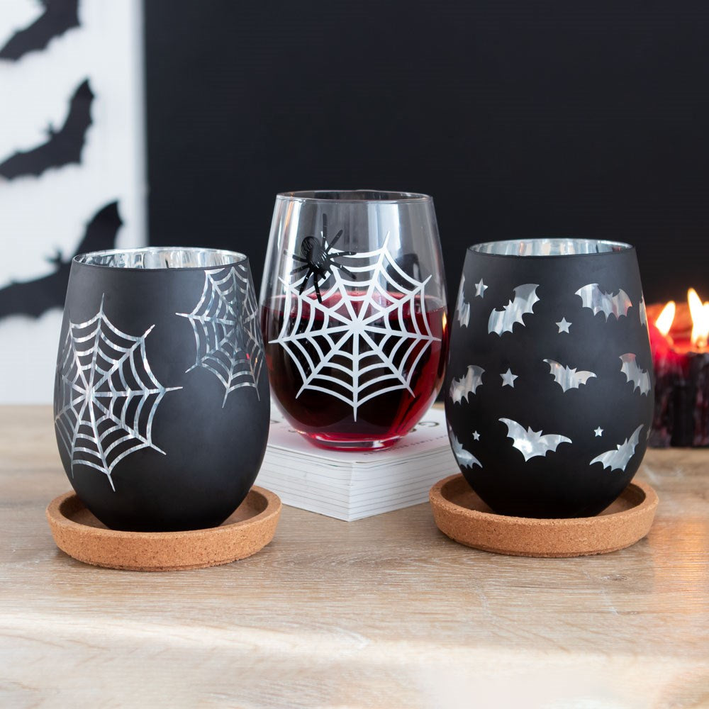 Three Halloween-themed glasses with spider webs, bats, and a spider on a wooden table.