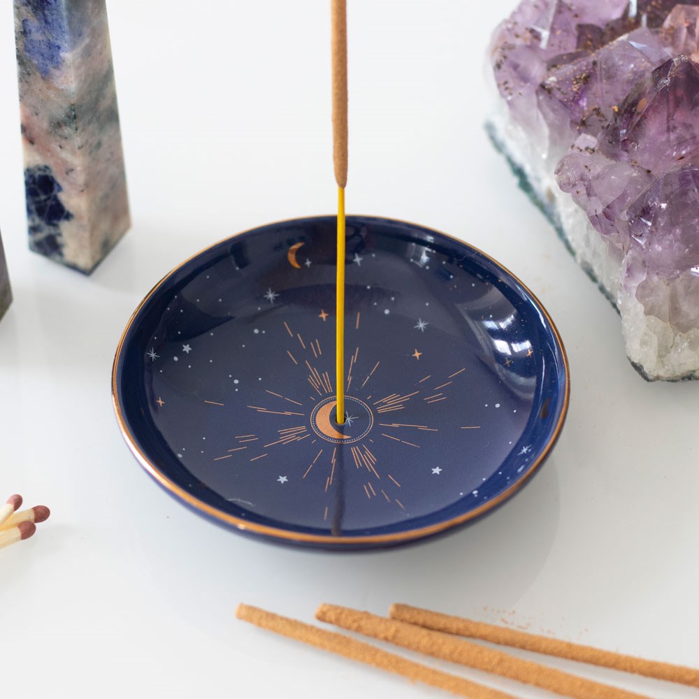 Incense stick burning on a blue dish with celestial design, surrounded by crystals on a white surface.