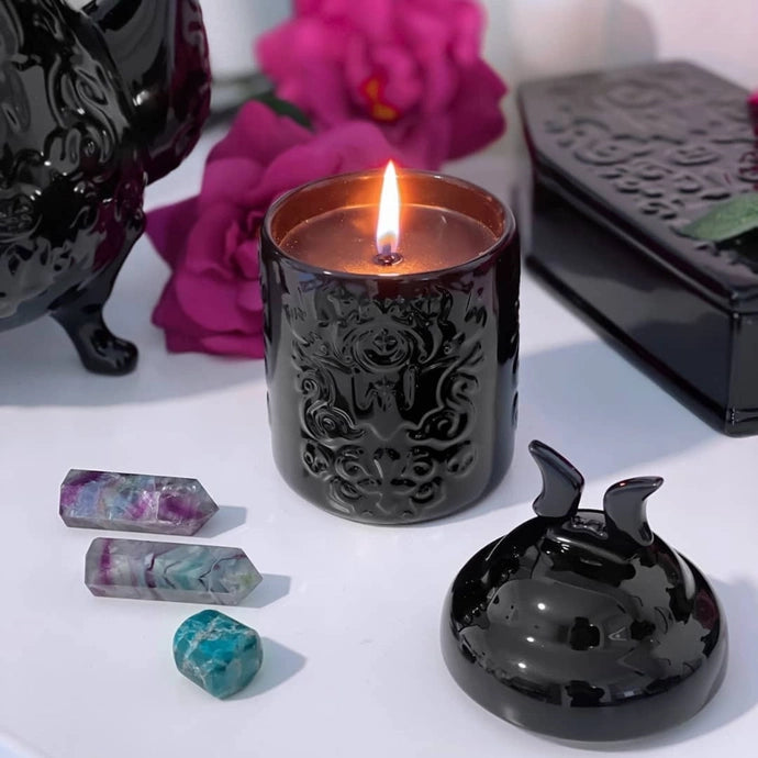 Decorative black candle with intricate designs, surrounded by purple flowers and decorative stones on a white surface.